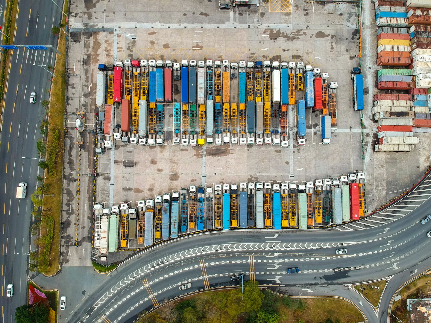 Many semi-trucks and stacked shipping containers fill a large logistics yard.